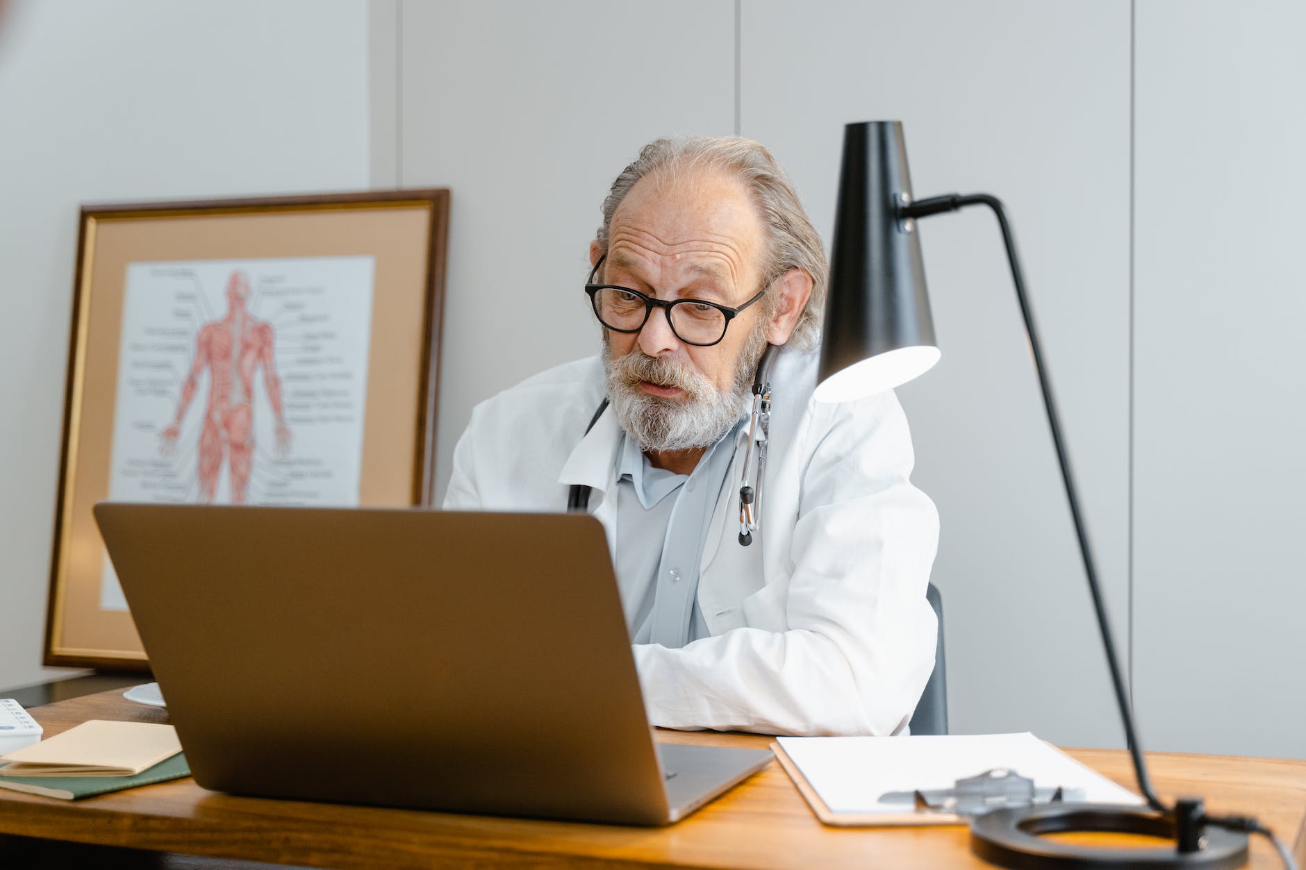 an elderly man in white lab coat talking while facing the laptop